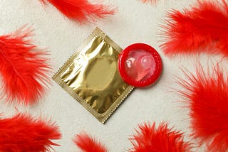 A gold foil condom wrapper lies on a light textured surface next to an unrolled red condom. Surrounding the items are several bright red feathers, adding a soft, vibrant contrast to the scene. The lighting is even, and the image has a clean, playful feel.