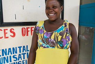 A pregnant woman wearing a yellow dress smiles outside of the door to the clinic. The sign behind her reads: Services Offered: OPD, Antenatal, Immunization, HIV Counseling.