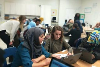 School classroom full of students seated at different tables. In the foreground there is a medium skin toned woman in a blue shirt and grey headscarf to the left, a light skin toned young girl with curly hair and a brown shirt in the middle and a dark skin toned girl with a black puffer coat and a blue, green and yellow headscarf to the right. The girl in the middle is on a laptop showing something to the girl on the left. The girl on the right is on her own laptop.