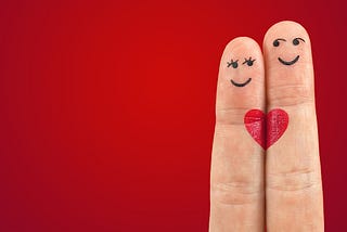 Two fingers close together, the shorter one has female eyes and mouth drawn on it, the taller has male eyes and a smile drawn on it, Each has half a red heart which — side by side — make a whole heart