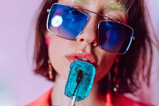 A person wearing large blue sunglasses holding a blue lollipop containing a scorpion up to their lips.