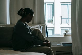 Black woman sitting on a bed beside a window while deep in thought