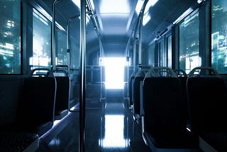 This image depicts the interior of a bus, with a futuristic or moody atmosphere. The lighting is dim, with a strong bluish tone dominating the scene. The seats are empty, and the bus appears to be traveling through a city at night, as suggested by the faint city lights visible through the windows. The light at the end of the bus creates a dramatic effect, emphasizing a sense of mystery or solitude. The reflective floor adds to the overall sleek, modern feel of the scene.