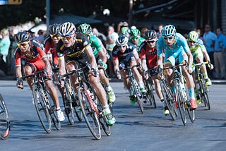 Competing bicyclists cooperate to form a peloton. Photo by Quino Al on Unsplash. Image shows a dozen bicyclists riding close together.