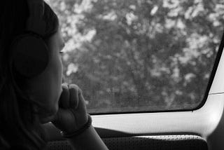 ID: Black and white photo of a long-haired person in profile. They’re looking out of a car window and wearing headphones.