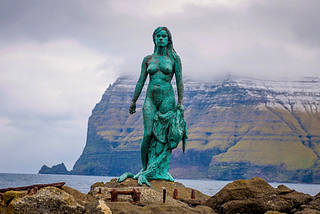 Photo of bronze sculpture of Selkie Seal-Woman standing on a rock in a bay in The Faroe Islands.