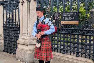 A man dressed in a red tartan kilt playing the bagpipes against black railings