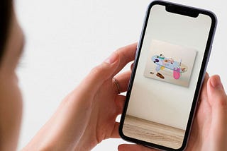 A woman holds a phone. She’s trying out a piece of art on her walls using Augmented Reality, and seeing how it would look if she bought it.