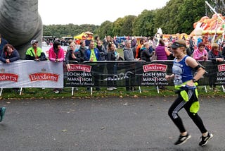 A blurry image of a woman in a blue vest and black leggings smiling as she runs past a crowd.