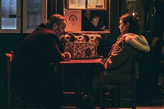A man and woman sit at a table facing each other with serious, sad facial expressions.
