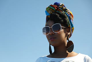 J LeShaé pictured from the shoulders up in shades and an African print head wrap against the sky. Photographer: Krystal René.