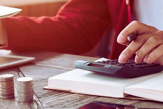 Na zdjęciu znajduje się osoba siedząca przy drewnianym stole, obliczająca coś za pomocą kalkulatora. Na stole leżą stosy monet, otwarty notes, tablet oraz dokumenty. W tle widać dłoń trzymającą rachunek, co sugeruje, że osoba ta zajmuje się budżetowaniem, obliczeniami finansowymi lub planowaniem wydatków. Zdjęcie przekazuje poczucie koncentracji i troski o finanse, a także zachęca do podejmowania świadomych decyzji finansowych.