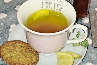 A cup of herbal tea with a slice of lemon and a biscuit on the saucer. The cup has the words ‘Always Time For Tea’ inscribed at the top.