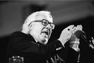 A black-and-white photograph of Ray Bradbury talking at the Miami Book Fair International in 1990. He has white hair and glasses with black frames. He is holding his hands up while talking into a microphone. He has a ring with a dark jewel on one finger.