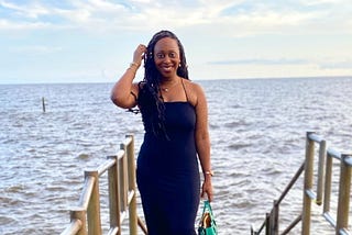 Picture of brown skinned black woman smiling. She is wearing a long black sleeveless dress and is standing on the dock with river behind her. The sky is clear with a few clouds.