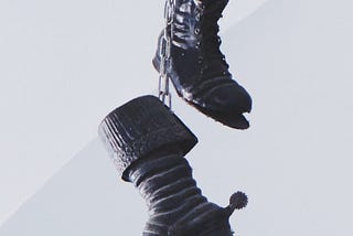Two worn-out black boots hang from chains against a greyish background