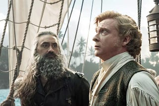 Taika Waititi as Blackbeard looks at Rhys Darby as Stede Bonnet, while Stede looks away with a fearful expression. Blackbeard is in all leather and Stede is wearing an 18th-century outfit with a white blouse and vest. They are on a ship.