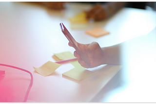 Photograph showing a person using a smart phone in a meeting room with others present.