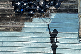 Picture of a bunch of old 33 rpm records nailed to a wall, to look like balloons, with string attached to each and bundled to the bottom, of a silhoutte of a girl painted on the wall, which looks like the balloons are carrying the girl away.