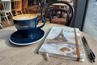 Photo of a cup of coffee, a journal, and a pen on a table in a coffee shop.