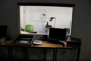 A usability lab setup, separated by glass. On one side, a researcher sits, observing the user, who sits on the other side.