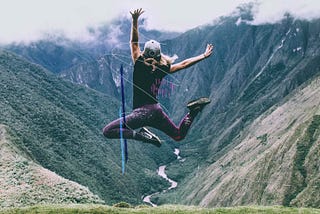 Person jumping high in front of a mountain scene wearing an athletic outfit and hat. They have blonde hair and appear to be caucasian.