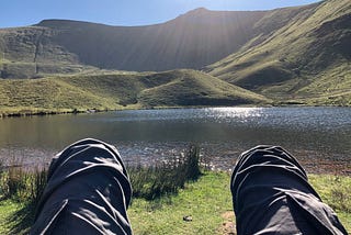 Man relaxed and looking over knees at a lake and hills in the sunshine.