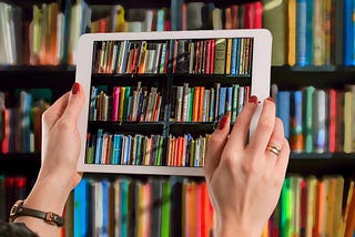 A person is holding up a tablet in front of a bookcase filled with books. They are taking a picture with the tablet, so the books are visible on the tablet screen as well. Only the person’s hands are visible. They wear read fingernail polish and have a ring on their right ring finger. They also wear a watch on their left wrist.