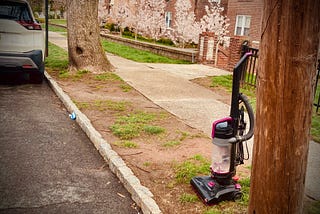 Image shows a curb between a street and tree-lined sidewalk. An abandoned upright vacuum rests there, awaiting garbage collection.