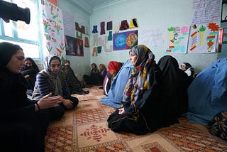 A group of women wearing colourful head scarves sit on the floor in conversation.