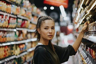 Mulher de cabelo castanho escuro e preso com uma blusa preta no corredor do mercado.