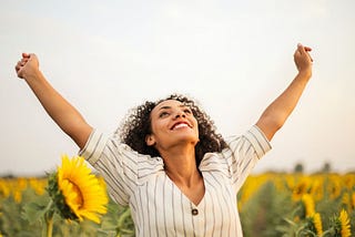 A woman standing in a field of sunflowers raises her arms to the sky