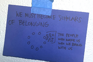 Black handwriting on blue paper, taped to a white wall with a piece of blue masking tape. The text is “WE MUST BECOME SCHOLARS OF BELONGING” and next to a doodle of circles meant to depict people sitting in a circle, one of them with a cloud of people behind them labeled, “THE PEOPLE WHO MAKE US WHO WE BRING WITH US”