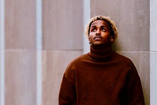 A man in a brown sweater stands by a gray stone wall and looks up at the sky