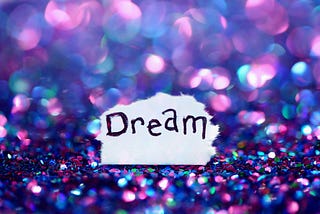 Purple, pink, and blue sparkling gems with a torn corner of the paper in the center. The word “dream” is written on the paper.