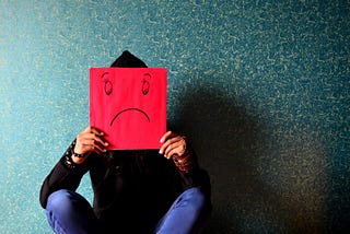 A person sitting in front an interior wall, holding a red sign with a sad smiley in front of their face.