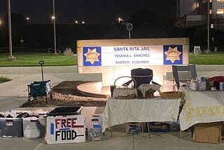 Two tables and some bins are set up on a wide walkway in front of an official sign saying “Santa Rita Jail” with the name of Sheriff Yesenia L. Sanchez. On the tables are baskets of food and containers of drinks. The bins are full of items with an attached homemade sign that says “FREE FOOD.” A few lights shine in the night sky in the background.