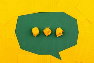 On a green table, pieces of yellow lined paper are arranged in an oval to create a speech bubble, which contains three balls of scrunched-up yellow paper to indicate conversation.