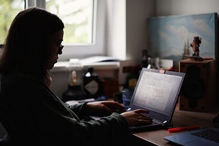 Woman sitting at desk using laptop. Window on left side lets in bright sunlight so woman is mostly in silhouette.