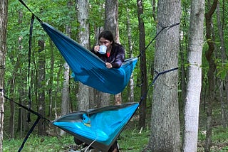 Kaitlyn sits in the top hammock of a stack of two, sipping coffee from an IBM branded mug and gripping the fabric with her free hand, as the dog and person in the lower hammock are being flipped out onto the forest floor. (Dog and human are fine and still attempting crazy stunts, today.)