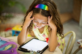 Girl wearing a rainbow headband sitting and looking and down at a tablet device.