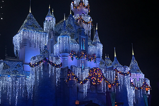 Night shot of Sleeping Beauty Castle at the Disneyland Resort decorated for the holidays with white icicle lights, multi-color lit garland, and a wreath.