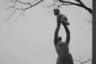 -the “joy of life” sculpture by czech sculptor jan hàna, donated to japan by czechoslovakia socialist republic in 1980 as peace symbol, in commemoration of the nagasaki atomic bomb, is a sculpture of a jubilant mother holding up her baby. it was made of bronze and the colour is ash-grey. the photograph itself is the writer’s personal collection, taken during a trip to nagasaki peace park in spring 2018.