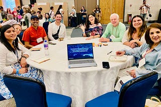 Eight people sit at a roundtable with an open laptop.