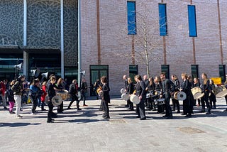 Two groups of percussionists face each other, one group carrying tall speakers on their backs, outdoors in front of a Theatre.