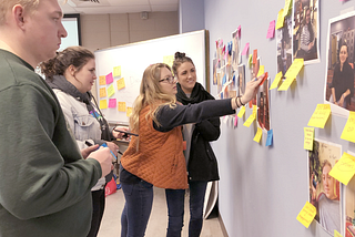 Four students place photographs and sticky note comments on a classroom wall