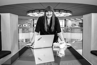 A woman with long straight hair and a fringe stands at a table and smiles, an open laptop is on the table in front of her.