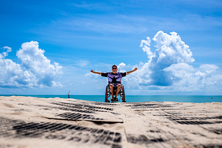 Person in wheel chair on the sand with arms outstretched. Bright blue ocean and clouds in the background.
