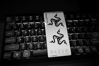 A sheet of Razer’s famous stickers sitting on top of a powered-on Keychron V1 gaming keyboard. The top two stickers depict the Razer snake logo and the bottom one is just their name. The photo is in black and white.