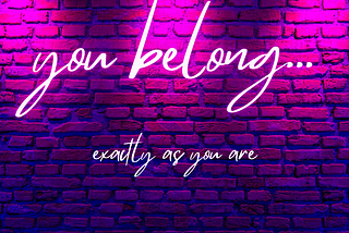 Image of a pink, purple, and blue lit brick wall with neon text reading “you belong… exactly as you are”